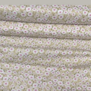 May include: A bolt of fabric featuring a repeating pattern of small white daisies with pink centers and green leaves on a light beige background. The fabric is tightly rolled, showing multiple layers of the floral print and a white paper label.