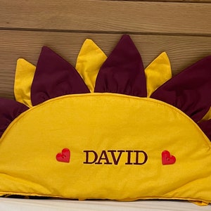 Può includere: Un cuscino a forma di sole giallo e bordeaux con il nome "DAVID" ricamato in nero e due bottoni rossi a forma di cuore.