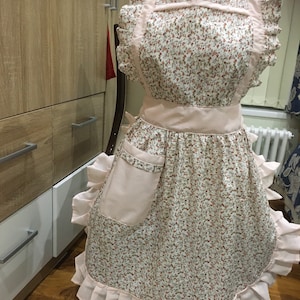 Op de afbeelding: Een roze en witte bloemenprint schort met een geruite rand en een grote zak. Het schort heeft een roze tailleband en een roze nekband.