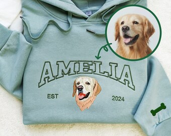 May include: A light blue hooded sweatshirt with the name "Amelia" embroidered in green on the front. The sweatshirt also features a small embroidered dog and the text "EST 2024". A bone is embroidered on the left sleeve.