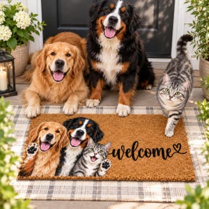 Peut inclure: Un paillasson rectangulaire en fibre de coco marron avec une impression représentant un golden retriever, un bouvier bernois et un chat tigré qui font signe de la patte. Le mot "welcome" est écrit en noir avec un petit cœur sur la droite.