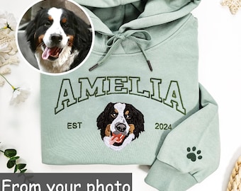 May include: A light green hoodie with the name "AMELIA" embroidered in dark green. The hoodie features a dog portrait and paw print. The text "EST 2024" is also embroidered on the hoodie. The text "From your photo" is at the bottom.
