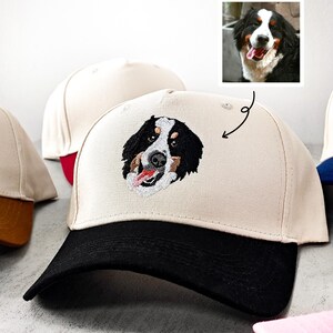 May include: A beige baseball cap with a black peak displays an embroidered Bernese Mountain Dog face. The dog's face is black, white, and brown, with a red tongue. Other caps are visible in the background.