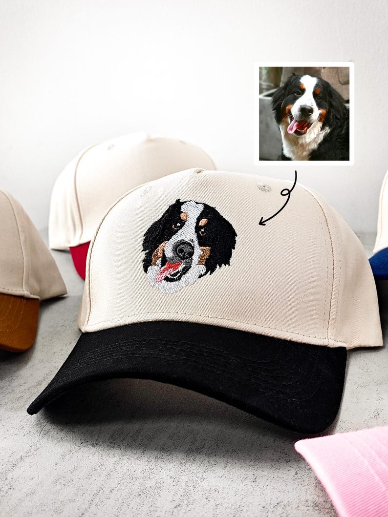 May include: A beige baseball cap with a black peak displays an embroidered Bernese Mountain Dog face. The dog's face is black, white, and brown, with a red tongue. Other caps are visible in the background.