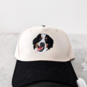 May include: A baseball cap with a cream-coloured crown and a black brim. The cap features an embroidered image of a Bernese Mountain Dog's face, showcasing black, brown, and white fur, with a red tongue. The hat is a classic six-panel design.
