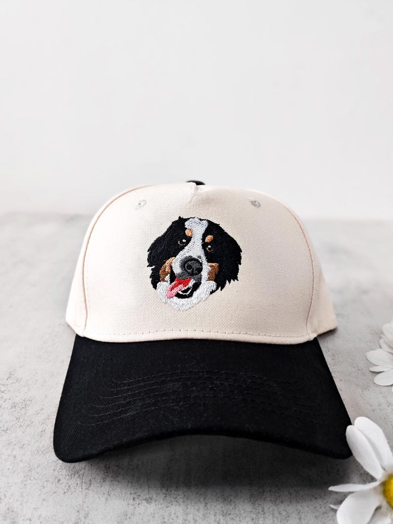 May include: A baseball cap with a cream-coloured crown and a black brim. The cap features an embroidered image of a Bernese Mountain Dog's face, showcasing black, brown, and white fur, with a red tongue. The hat is a classic six-panel design.