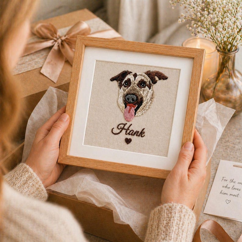 May include: A framed embroidered portrait of a dog with brown and black markings, a pink tongue, and the name "Hank" in cursive. The artwork is in a wooden frame with a white mat. A small heart is below the name.