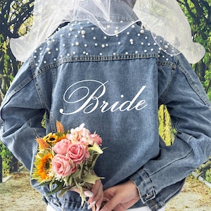 May include: A blue denim jacket with white pearl embellishments on the shoulders and the word "Bride" embroidered on the back. The jacket is worn by a person holding a bouquet of pink and yellow flowers.