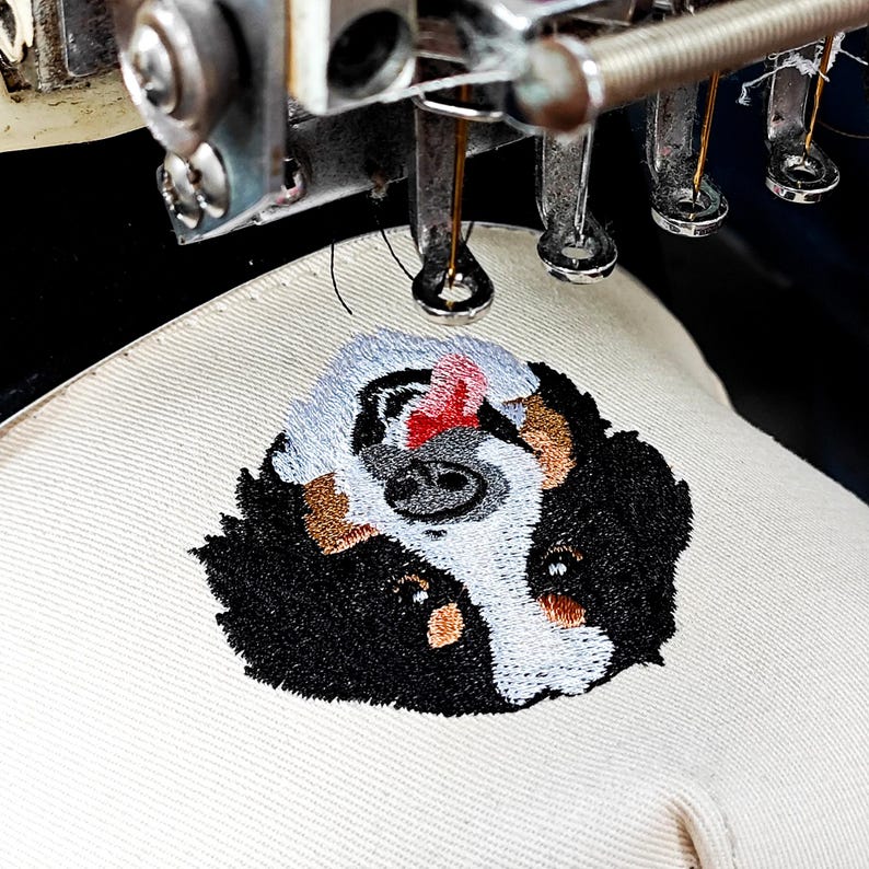 May include: An embroidery machine in action, creating a detailed dog face design on beige fabric. The dog's face features black, white, and brown colours, with a red tongue. The machine's needle is visible, adding intricate details.