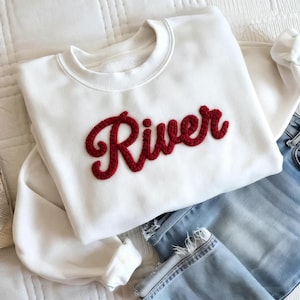 Può includere: Felpa bianca con la scritta "River" ricamata in rosso sul petto. La felpa è abbinata a jeans di denim blu chiaro. L'immagine è scattata con luce naturale.