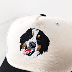 May include: A baseball cap with an embroidered Bernese Mountain Dog design. The cap has a cream-coloured crown and a black peak. The dog's face is detailed with black, white, brown, and red thread.