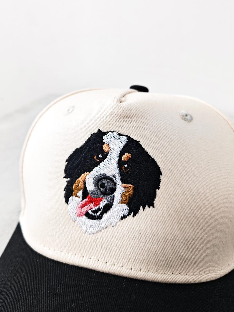 May include: A baseball cap with an embroidered Bernese Mountain Dog design. The cap has a cream-coloured crown and a black peak. The dog's face is detailed with black, white, brown, and red thread.