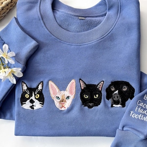 Può includere: Una felpa blu pervinca con facce di animali domestici ricamate: un gatto bianco e nero, un gatto senza peli, un gatto nero e un cane. La manica ha il testo "Bay Cactus Hades Toothless".