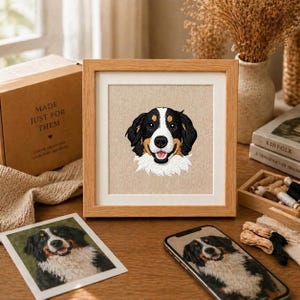 May include: A framed embroidery of a Bernese Mountain Dog with a happy expression. The dog has black, white, and brown fur. The frame is light brown, and the background is beige. A box with the text "MADE JUST FOR THEM" is visible.