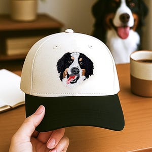 May include: A cream-coloured baseball cap with a black peak, embroidered with a Bernese Mountain Dog's face. The dog's tongue is visible. The cap is on a wooden surface.