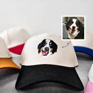 May include: A cream-coloured baseball cap with a black peak displays an embroidered Bernese Mountain Dog's face. The dog's tongue is visible. Other caps with different coloured peaks are in the background.