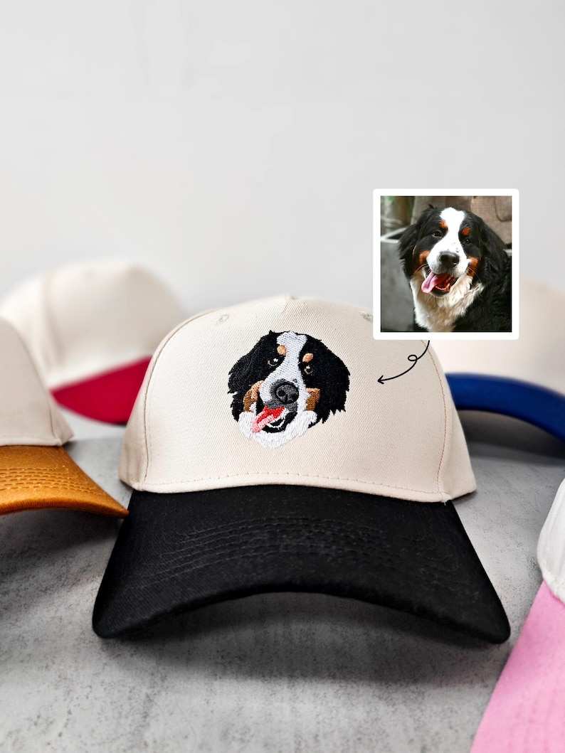 May include: A cream-coloured baseball cap with a black peak displays an embroidered Bernese Mountain Dog's face. The dog's tongue is visible. Other caps with different coloured peaks are in the background.