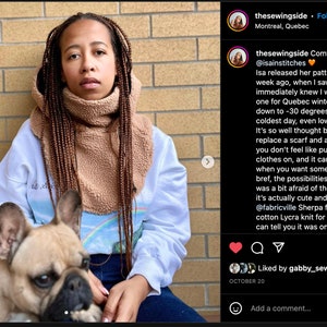 May include: A person wearing a white sweatshirt with a rainbow graphic and a brown fleece neck warmer. The person is holding a light brown French bulldog puppy. The text "Comfort hood by @isainstitches" is visible in the image.
