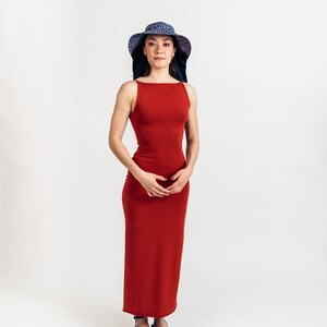 May include: A woman wearing a sleeveless, floor-length, rust-colored dress and a wide-brimmed hat with a blue and purple pattern. She is also wearing black sandals. The background is plain white.