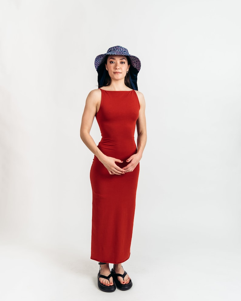 May include: A woman wearing a sleeveless, floor-length, rust-colored dress and a wide-brimmed hat with a blue and purple pattern. She is also wearing black sandals. The background is plain white.
