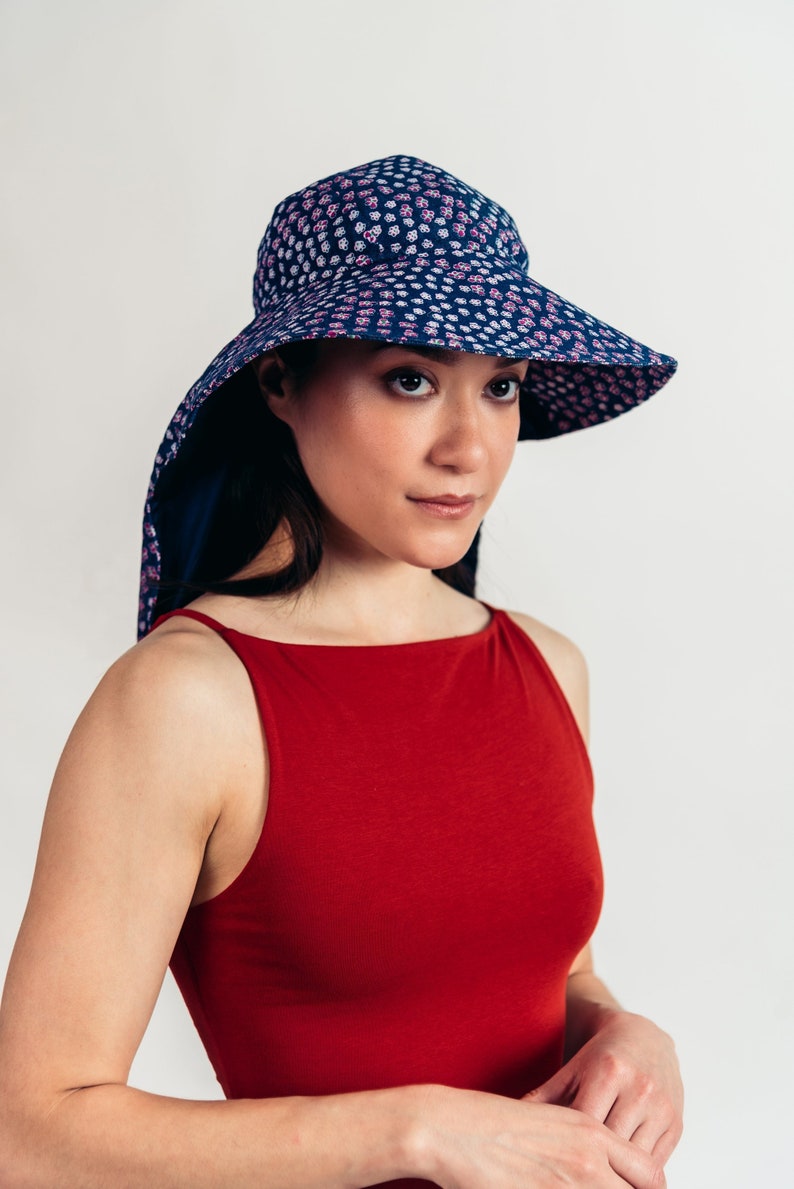 May include: A navy blue sun hat with a wide brim and a neck drape, patterned with small white and pink flowers. The hat is worn with a red tank top. The hat provides sun protection.