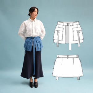 Op de afbeelding: Een blauwe utility-rok met een tailleband inclusief een lange riem, twee grote cargozakken aan de voorzijde met klepsluiting en verticale banddetails. Het ontwerp wordt vergezeld door twee technische lijntekeningen van voor- en achteraanzicht.