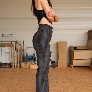 Puede incluir: Una mujer con un top negro corto y pantalones de cuadros de cintura alta. Est&aacute; de pie en un garaje con cajas en el fondo.