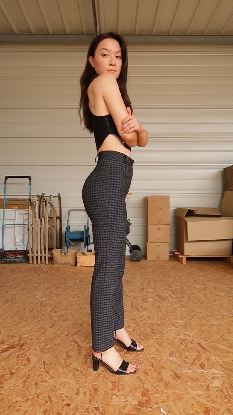 Puede incluir: Una mujer con un top negro corto y pantalones de cuadros de cintura alta. Est&aacute; de pie en un garaje con cajas en el fondo.