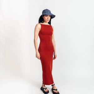 May include: A rust-colored, sleeveless maxi dress with a square neckline. The dress is paired with black sandals and a wide-brimmed sun hat with a blue and white polka dot pattern. The hat has a wide brim and a dark blue lining.