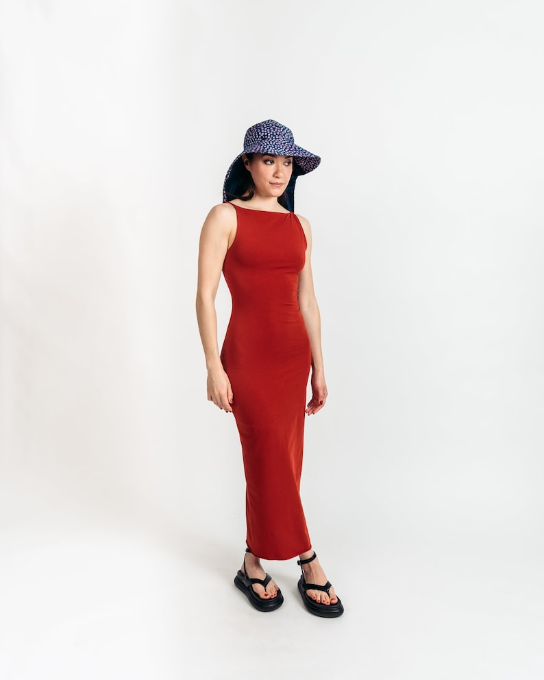 May include: A rust-colored, sleeveless maxi dress with a square neckline. The dress is paired with black sandals and a wide-brimmed sun hat with a blue and white polka dot pattern. The hat has a wide brim and a dark blue lining.