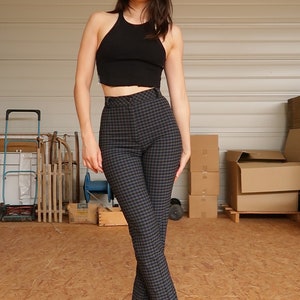 Puede incluir: Una mujer con un top negro sin mangas y pantalones de cuadros de cintura alta. Est&aacute; de pie en un almac&eacute;n con cajas y otros objetos en el fondo.