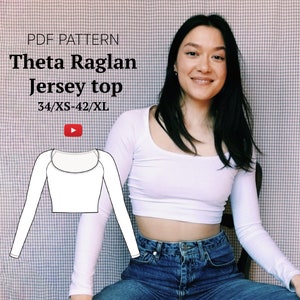May include: A white long-sleeved jersey top with a scoop neck and raglan sleeves. The top is cropped and is shown on a model wearing blue jeans. The text "Theta Raglan Jersey top 34/XS-42/XL" is displayed above the model.