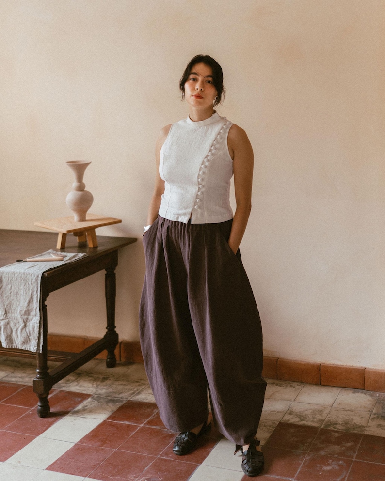 May include: A woman wearing a white sleeveless top with a high neckline and a row of white buttons down the side. She is wearing dark brown wide-leg pants and black shoes.