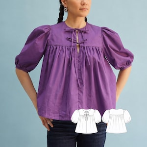 May include: A vibrant purple blouse featuring voluminous puff sleeves, a round neckline, and a front placket with two sets of thin fabric ties. The garment has a gathered bodice and includes a small white line drawing illustration of the design.
