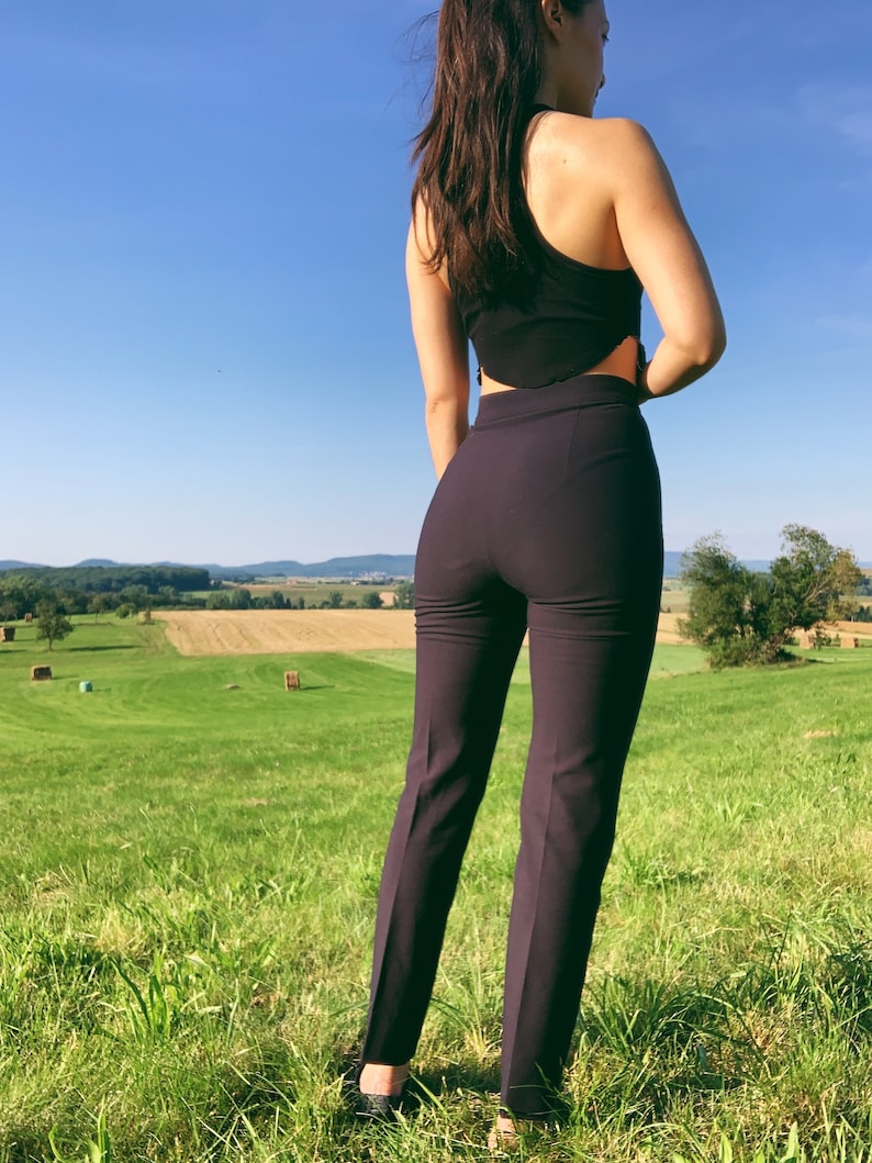 Puede incluir: Una mujer con un top negro corto y pantalones de color morado oscuro se encuentra de pie en un campo de hierba verde. El cielo es azul y hay balas de heno en la distancia.