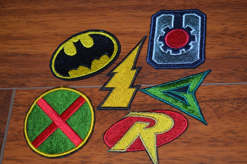 Op de afbeelding: Een set van zeven geborduurde strijkpatches met superheldenlogo's. De patches zijn in verschillende kleuren, waaronder rood, geel, groen en blauw. De logo's omvatten Batman, The Flash, Green Arrow, Robin en het logo van de Justice League.