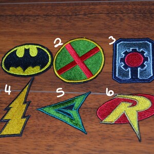 Op de afbeelding: Zes geborduurde patches met superheldenlogo's. De patches zijn in verschillende kleuren, waaronder zwart, geel, groen, rood, blauw en wit. De logo's omvatten Batman, The Atom, Cyborg, The Flash, Green Arrow en Robin.