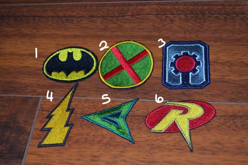 Op de afbeelding: Zes geborduurde patches met superheldenlogo's. De patches zijn in verschillende kleuren, waaronder zwart, geel, groen, rood, blauw en wit. De logo's omvatten Batman, The Atom, Cyborg, The Flash, Green Arrow en Robin.