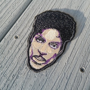 Op de afbeelding: Gestikte patch met een portret van Prince, een muzikant die bekend staat om zijn paarse heerschappij. De patch is zwart-wit met paarse accenten.