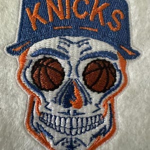 Peut inclure: Écusson brodé représentant un crâne portant un chapeau bleu avec le mot "KNICKS" en orange. Les yeux du crâne sont des ballons de basket, et le design incorpore des détails orange et blancs. Un accessoire unique sur le thème du sport.