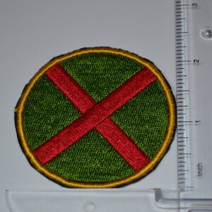 Op de afbeelding: Een ronde patch met een rode X op een groene achtergrond en een gele rand. De patch is ongeveer 5 cm in diameter.