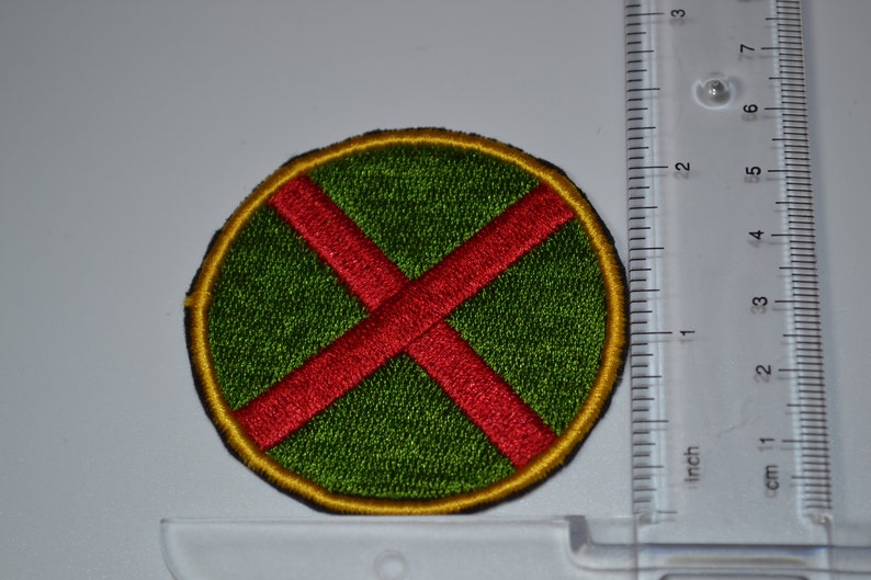 Op de afbeelding: Een ronde patch met een rode X op een groene achtergrond en een gele rand. De patch is ongeveer 5 cm in diameter.