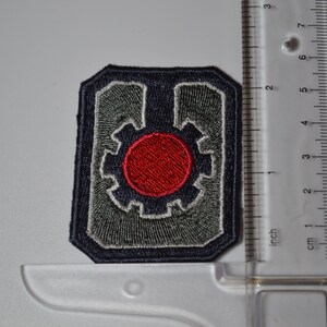 Op de afbeelding: Een geborduurde grijze en blauwe patch met een rode cirkel in het midden. De cirkel wordt omringd door een grijs tandwielachtig ontwerp. De patch is ongeveer 5 cm breed.