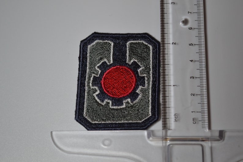 Op de afbeelding: Een geborduurde grijze en blauwe patch met een rode cirkel in het midden. De cirkel wordt omringd door een grijs tandwielachtig ontwerp. De patch is ongeveer 5 cm breed.