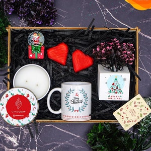 Puede incluir: Una caja de regalo navideña con un globo de nieve, bombones rojos en forma de corazón, una taza blanca con el texto "Merry Christmas", una vela y una pequeña planta en una maceta blanca. La caja está forrada con papel negro.
