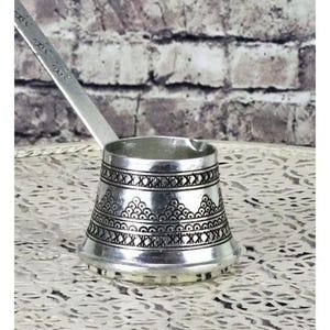 Puede incluir: Una cafetera turca plateada con un mango alargado. La cafetera presenta intrincados patrones geométricos repetidos alrededor de su cuerpo. El mango tiene texto grabado. La cafetera se encuentra sobre una superficie blanca estampada contra una pared de ladrillo.