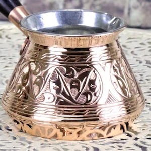 以下が含まれることがあります： 銀色の内装が施された、銅色の装飾が施されたトルココーヒーポット。ポットには複雑な彫刻模様と黒いハンドルが付いています。デザインは伝統的で、底が広く、上部が狭くなっています。