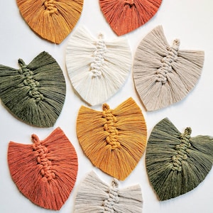 Op de afbeelding: Acht handgemaakte macramé bladwanddecoraties in verschillende kleuren, waaronder beige, wit, groen, oranje en geel. De bladeren zijn allemaal even groot en hebben dezelfde vorm, en zijn gemaakt van een dik, getextureerd garen.