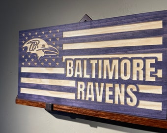 May include: A wooden sign with a purple stain and a distressed look. The sign features a carved American flag with the Baltimore Ravens logo and the words "Baltimore Ravens".