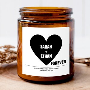 May include: A brown glass candle jar with a white label. The label has a black heart with the text "SARAH + ETHAN FOREVER" and the words "SLOWTIP GIFT CO. | VEGAN COCONUT SOY WAX HAND POURED WITH LOVE".